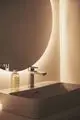 Waschtisch mit moderner Armatur von Ideal Standard, beleuchtet von einer runden Wandspiegel-Lampe. Flacher, rechteckiger Waschbecken mit fließendem Wasserhahn.